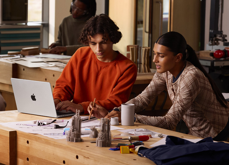 To personer samarbejder om et projekt ved et bord. Den ene skriver på en MacBook, mens den anden bruger blyant og lineal på papir, omgivet af tegneartikler.