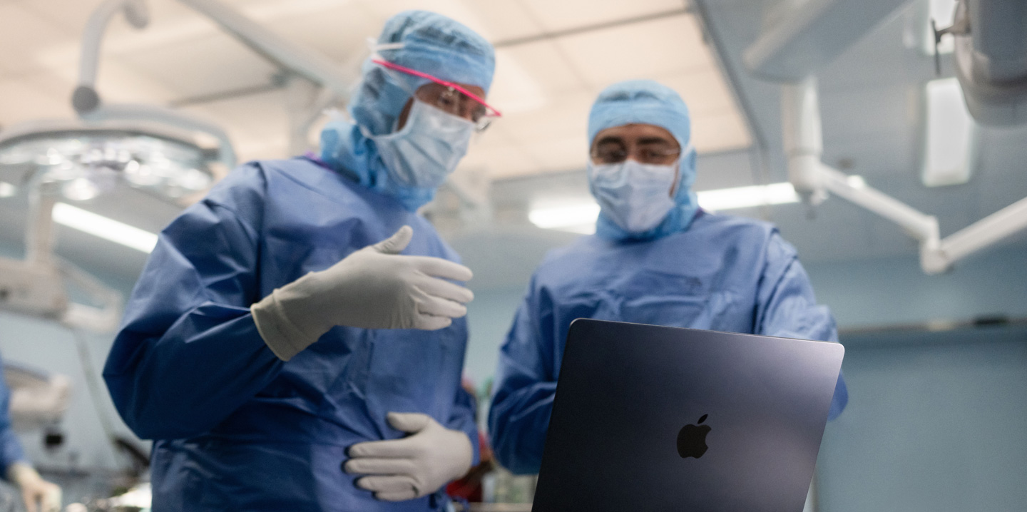 Dos cirujanos en el quirófano miran un MacBook, y uno hace gestos con la mano mientras el otro observa
