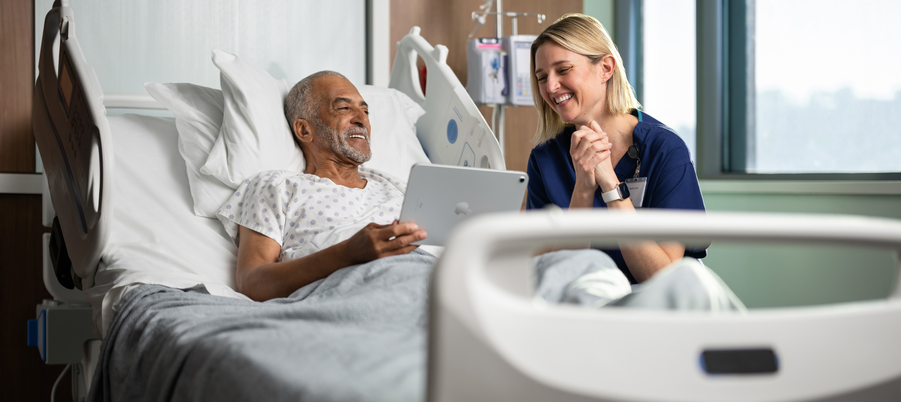 Un patient est assis dans un lit d’hôpital. Il regarde en souriant l’écran d’un iPad en compagnie d’une infirmière.
