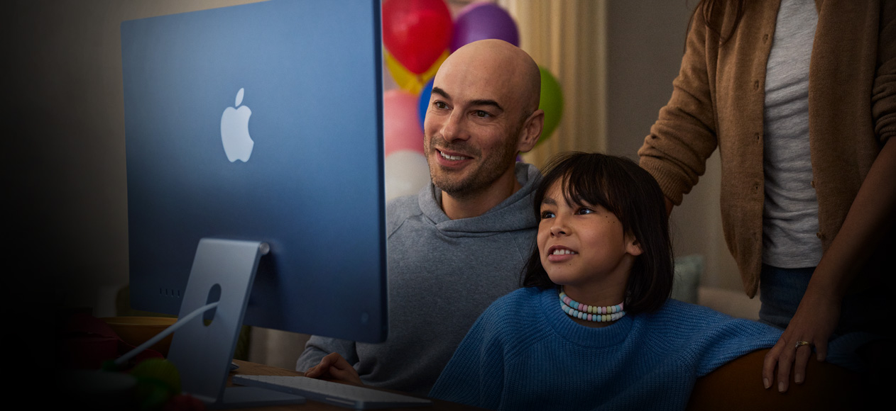 Két személy ül együtt egy iMac előtt, miközben egy harmadik mögöttük állva figyel.
